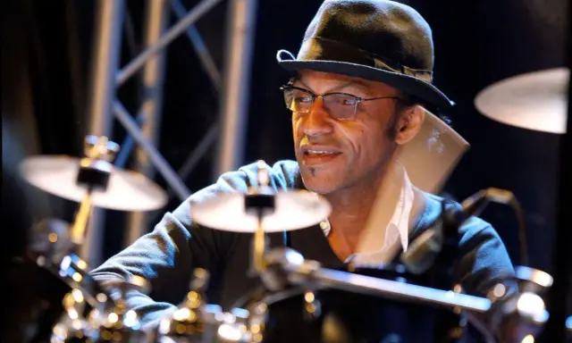 Un batteur concentré, portant un chapeau et des lunettes, joue sur scène. Il est difficile de dire combien de temps pour apprendre la batterie, mais il semble maîtriser son art.