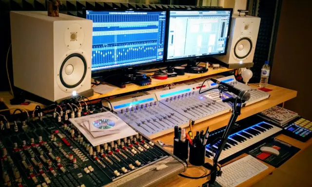 Studio d'enregistrement avec deux écrans affichant des pistes audio, des tables de mixage, des enceintes, un clavier MIDI et un microphone. Le fichier midi est prêt à être créé.