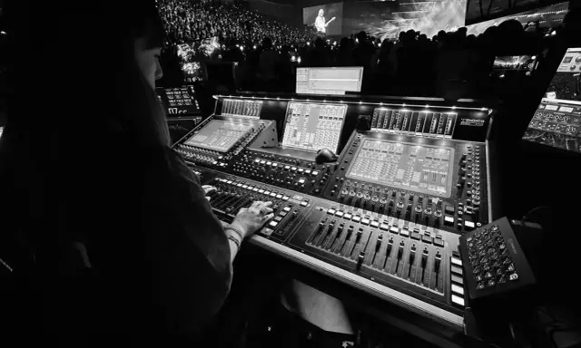 Une personne manipule une console de mixage dans un studio d'enregistrement, devant une foule lors d'un concert.