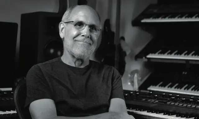 Dave Smith, souriant, dans son studio avec des synthétiseurs empilés.
