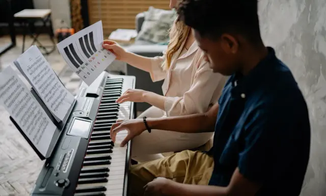 Deux jeunes jouent du piano, l'un tenant une partition montrant l'accord D7.