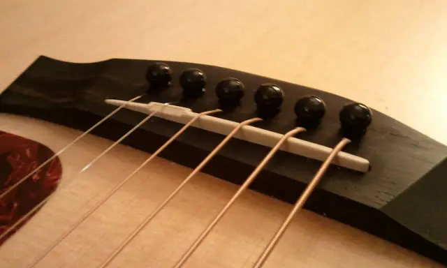 Gros plan sur le chevalet d'une guitare acoustique, avec ses six cordes prêtes à vibrer. Le bois sombre du chevalet contraste avec la pièce blanche où reposent les cordes.