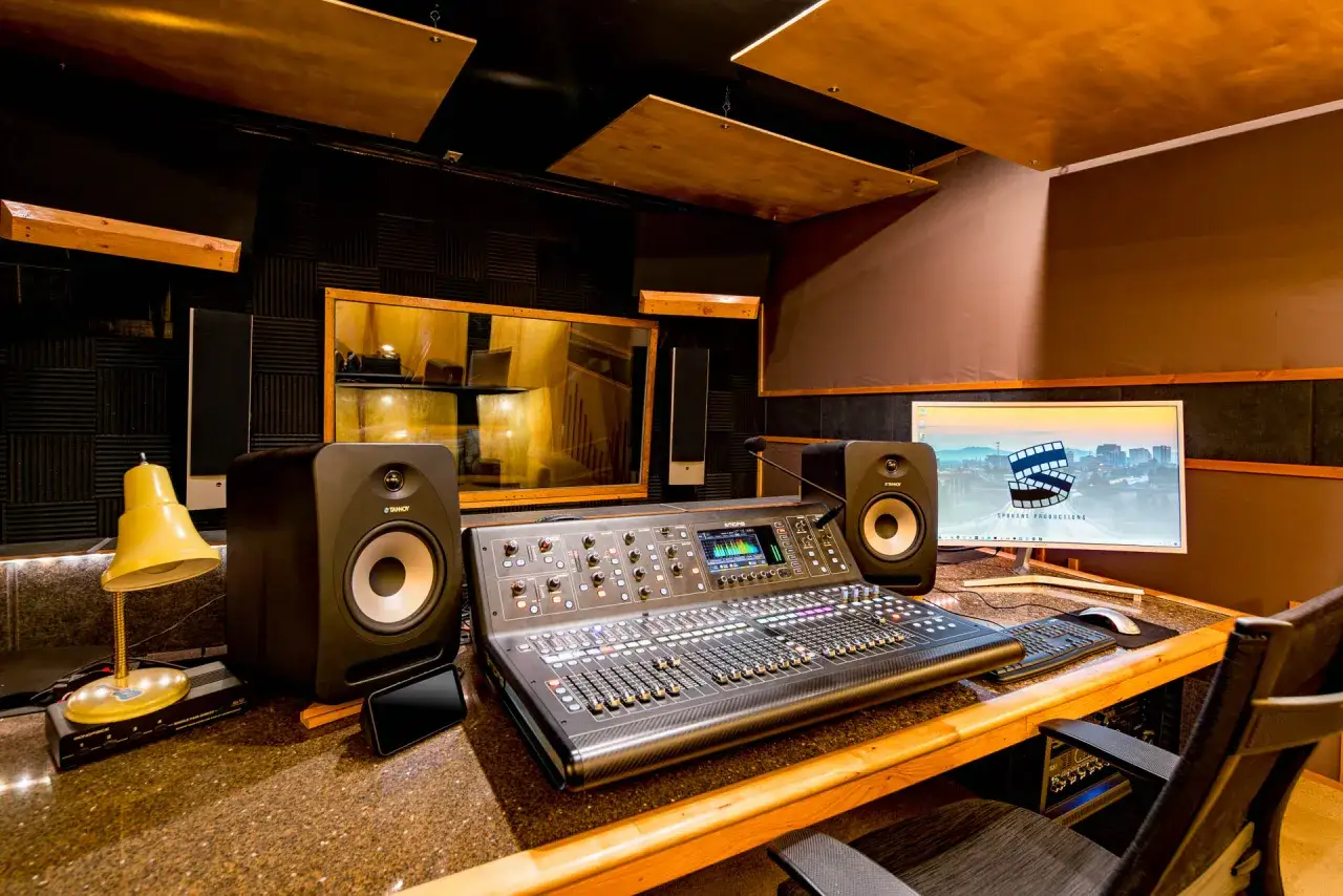 Console de mixage professionnelle dans un studio d'enregistrement, avec des moniteurs, un écran d'ordinateur affichant un logo de production et une lampe de bureau.