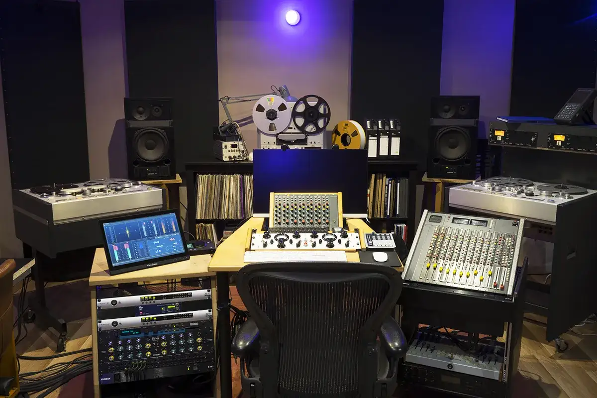 Studio d'enregistrement avec table de mixage, platines vinyles, magnétophone à bande et moniteurs. Le son analogique règne en maître.