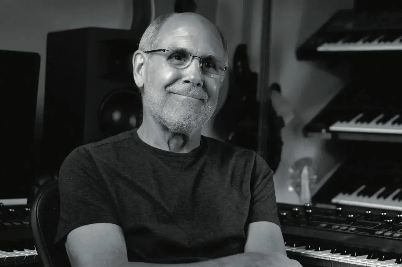 Dave Smith, le légendaire créateur de synthétiseurs, sourit dans son studio, entouré de ses instruments emblématiques.