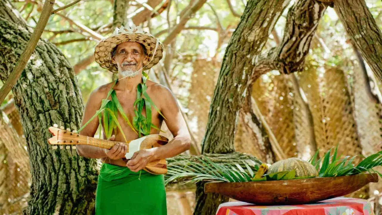 Un homme souriant, vêtu d'un pagne vert et d'un chapeau de paille, tient un ukulélé. Il évoque l'ukulélé origine, un instrument aux racines polynésiennes.