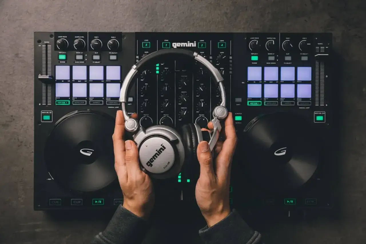 Mains d'un DJ tenant un casque argenté au-dessus d'une table de mixage Gemini, prêt à apprendre à mixer.
