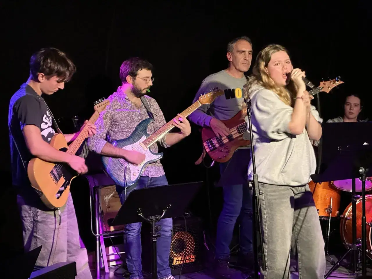 Un groupe de musique joue sur scène. La chanteuse, au premier plan, semble vouloir comment se faire remarquer.