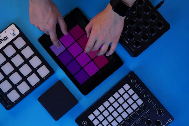 Des mains manipulent une tablette affichant des pads colorés, entourée de contrôleurs midi. Création musicale en cours.